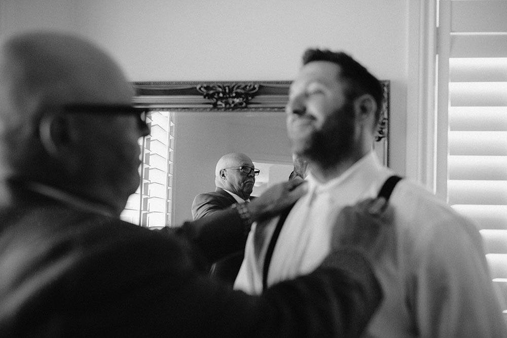 father helps his son fix his suspender straps in the mirror on his wedding day