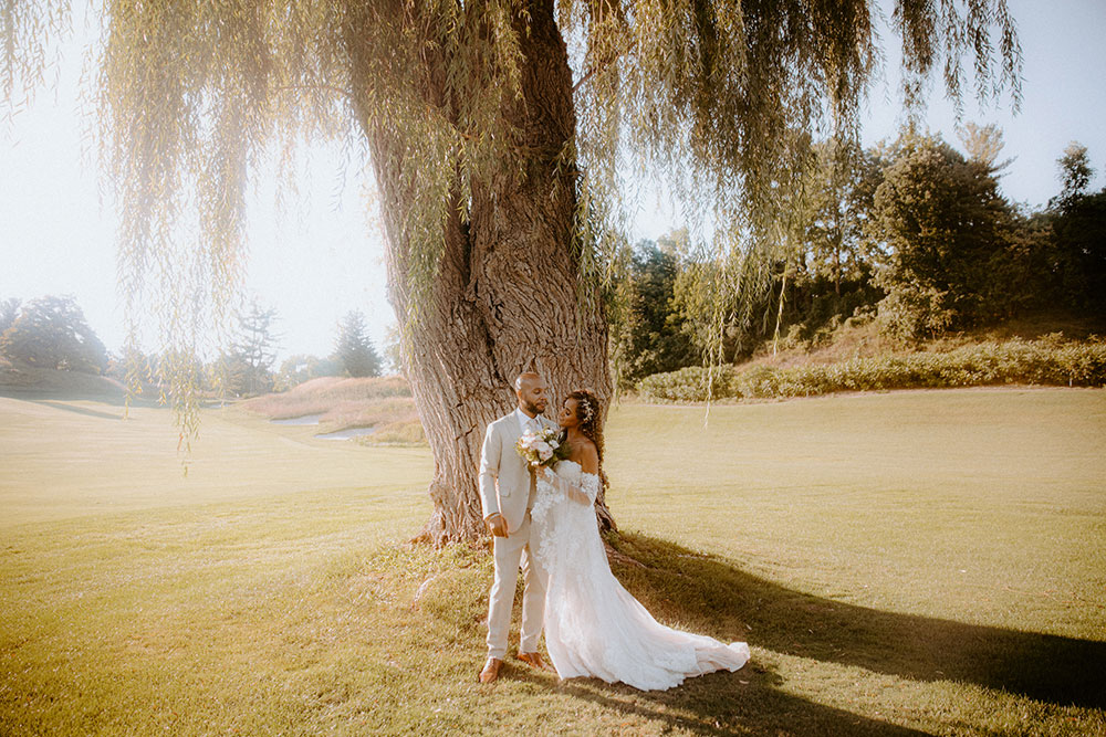 bride and groom pose by a giant willow tree at this Bayview Golf Wedding