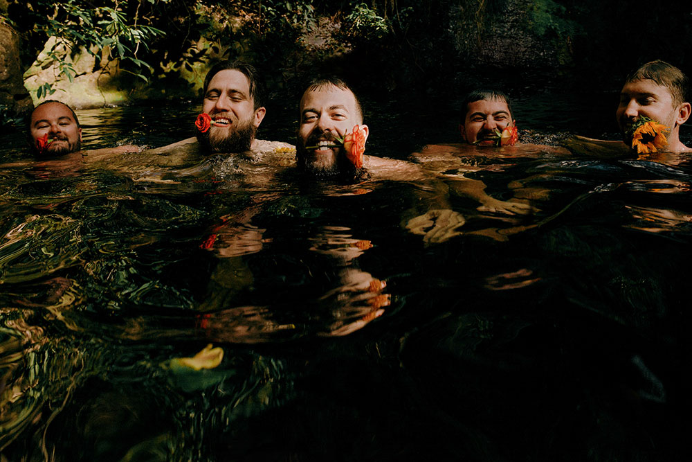 men with flowers in their mouth while they swim under a waterfall in the jungle at this belize wedding