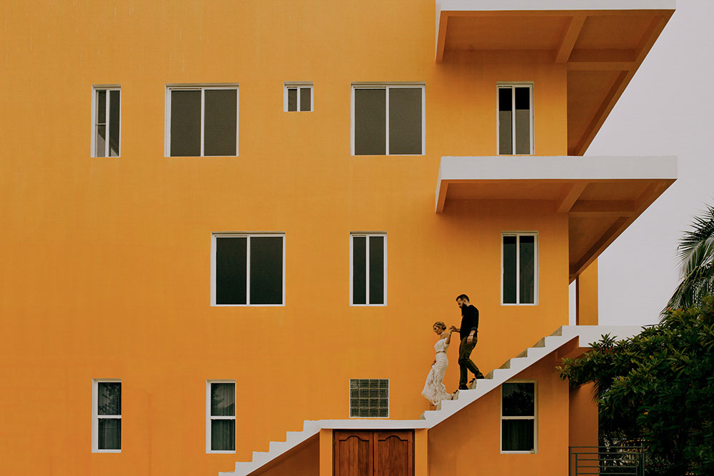 couple walk down stairs of a bright orange building in placencia at their belize wedding