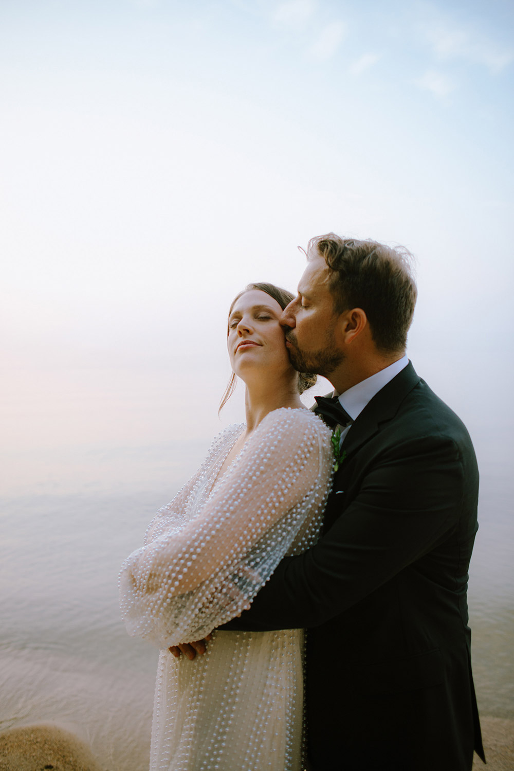 a soft kiss on the face from a groom to Bride as they embrace while taking their wedding portraits on the beach at the Northridge Inn in Muskoka