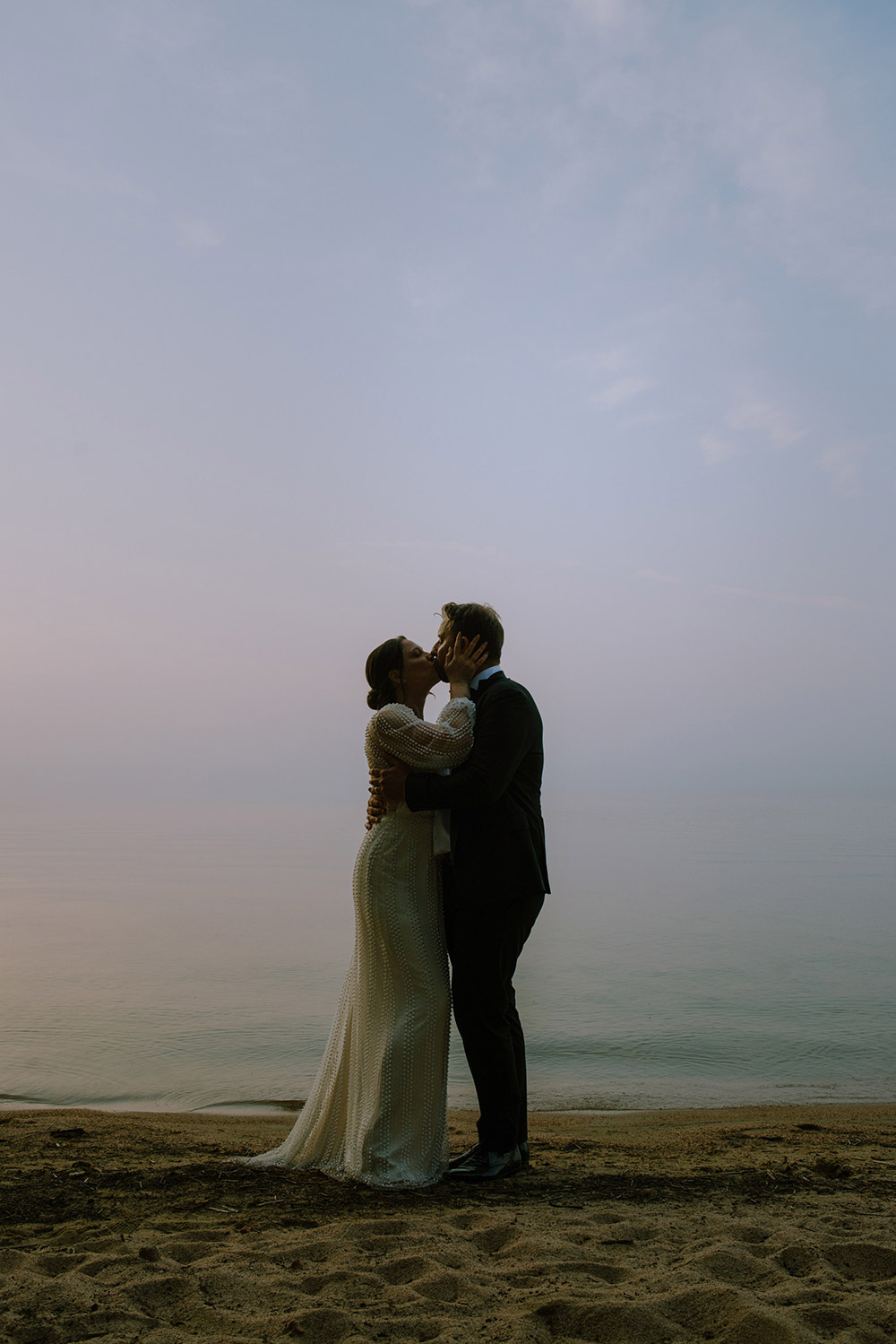 The bride and groom kiss and share a quiet moment as the sun sets on the sandy beach of he Northridge Inn resort which is located in thestunning Muskokas