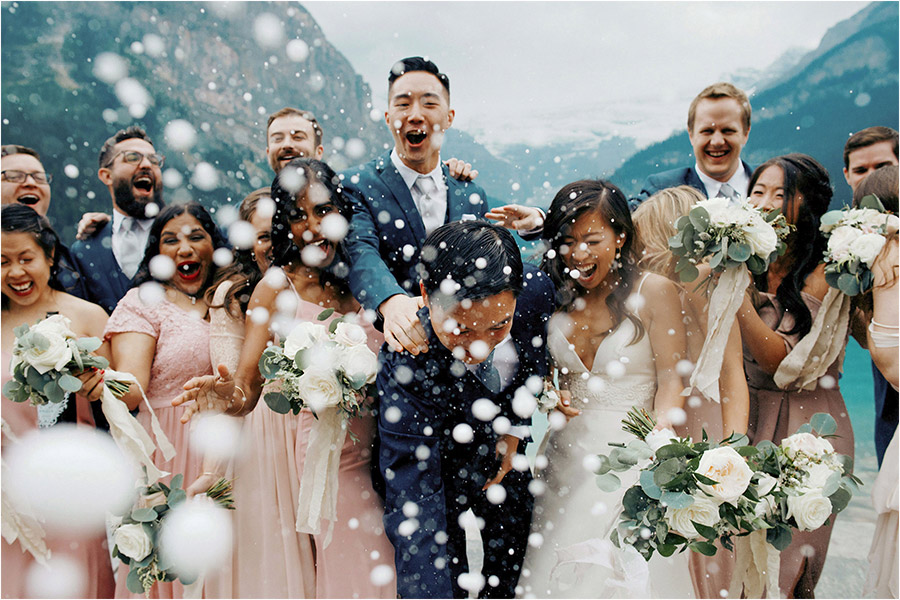 Lake Louise Wedding Photography of a wedding party popping a champagne bottle at the camera lens surrounded by the breathtaking lake louise mountains